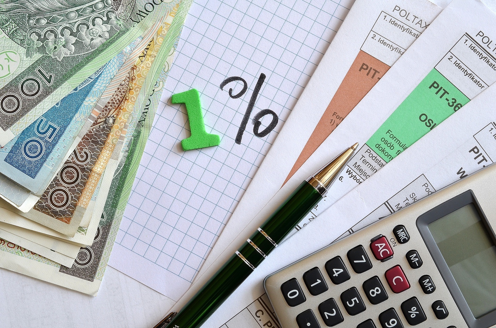 zdjęcie przedstawia dokumenty podatkowe, kalkulator i pieniądze lezące na stole, na środku na kartce została napisana informacja 1%