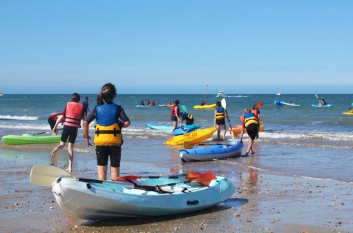 zdjęcie przedstawia dzieci na plaży, które niosąc kajaki wchodzą do może, dzieci są ubrane w kapoki