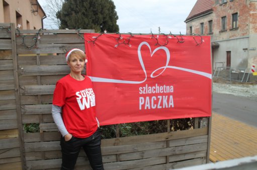 przed flagą akcji "Szlachetna paczka" stoi kobieta w czapce Świętego Mikołaja