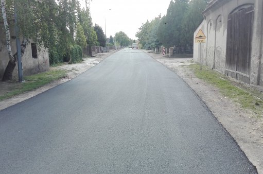 zdjęcie przedstawia prace nad nakładką asfaltową na jednej z gminnych dróg, widzimy świeży asfalt, po prawej i lewej stronie zabudowania
